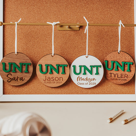 University of North Texas ornaments featuring green UNT letters with engraved personalization.  Shown in four wood types, from left to right, walnut, cherry, white and mahogany.  Officially licensed Mean Green personalized ornament by Half Turn Designs.