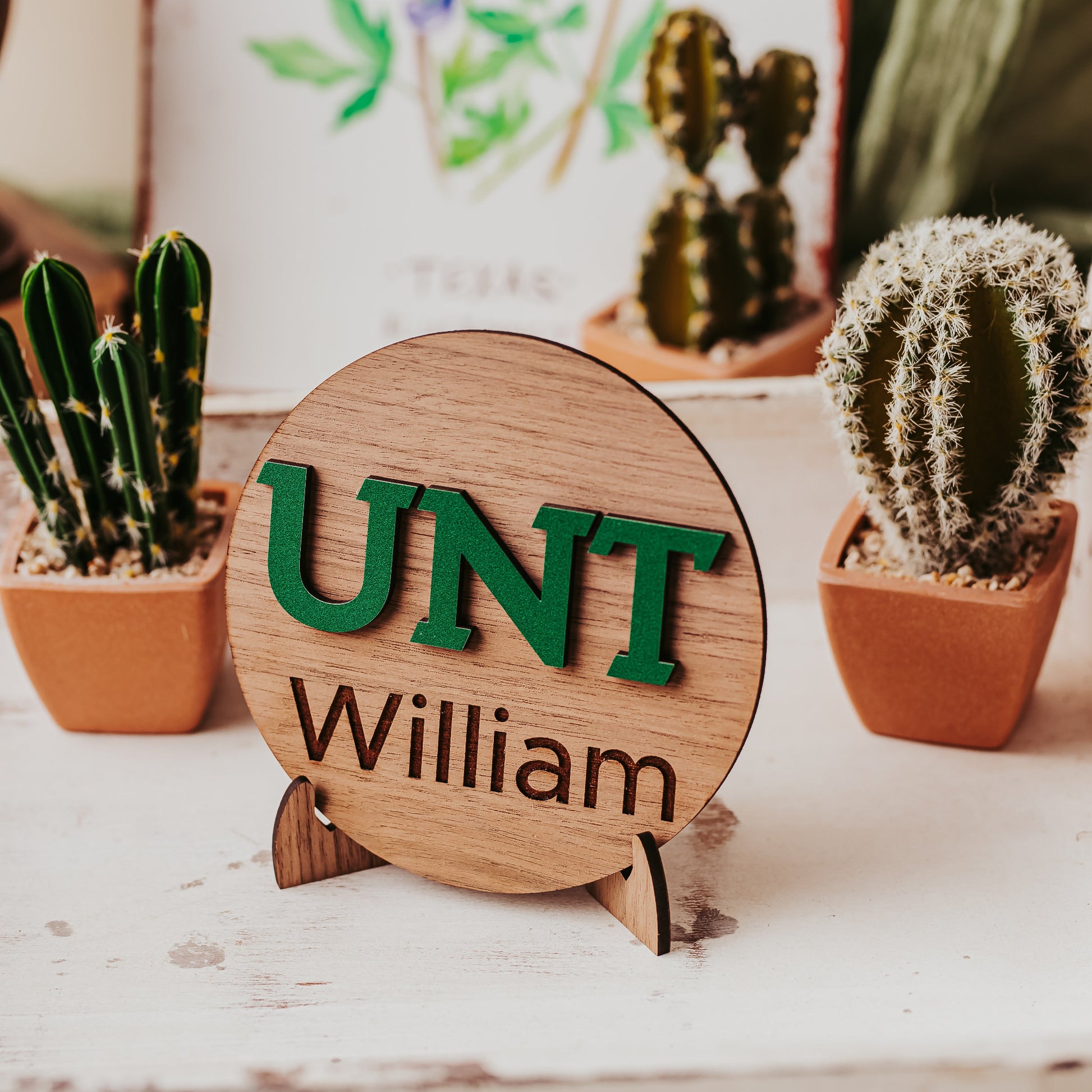 Officially licensed University of North Texas 5 inch round wooden dorm decor, shelf sitter and desk sign in walnut wood finish with the UNT letters in Mean Green, personalized with your choice of 1-2 lines of engraved text in four wood finishes. Comes with a matching stand.