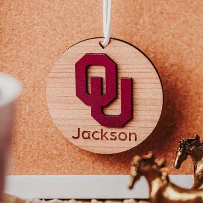 Engraved University of Oklahoma ornament featuring crimson OU logo on 3-inch round wood ornament, customizable with name or phrase, available in Cherry, Mahogany, Walnut, Sapele, or White finishes. Perfect Sooner Christmas gift or car charm. Shown here in Cherry wood.