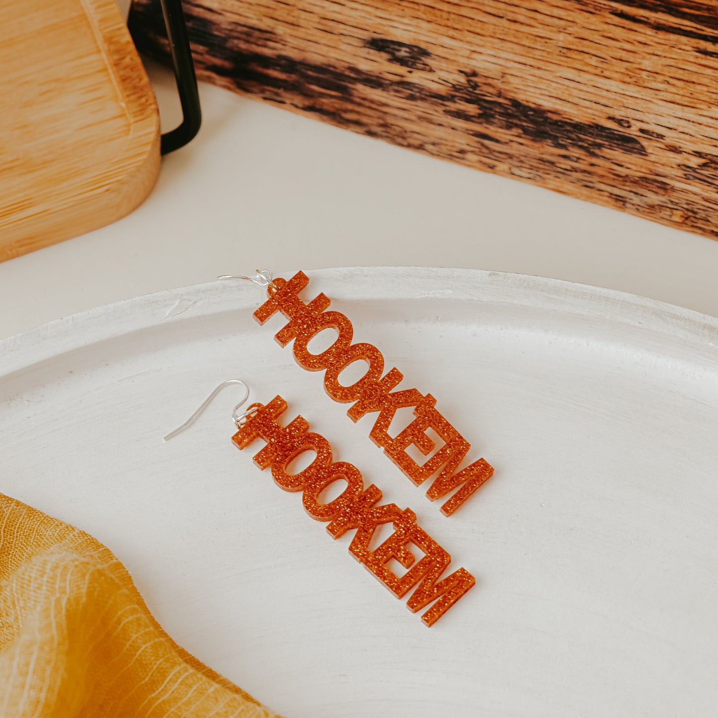 A pair of University of Texas Longhorns glitter dangle earrings featuring burnt orange acrylic word designs that spell “HOOK ’EM" on silver earring hooks