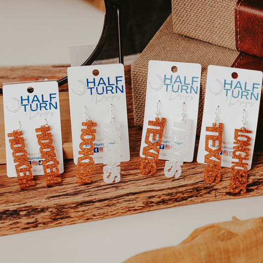 Four pairs of University of Texas Longhorns glitter dangle earrings on Half Turn Designs display cards, featuring burnt orange and white acrylic word designs that spell “HOOK ’EM,” "HORNS," “TEXAS,” and “LONGHORNS” on silver hooks
