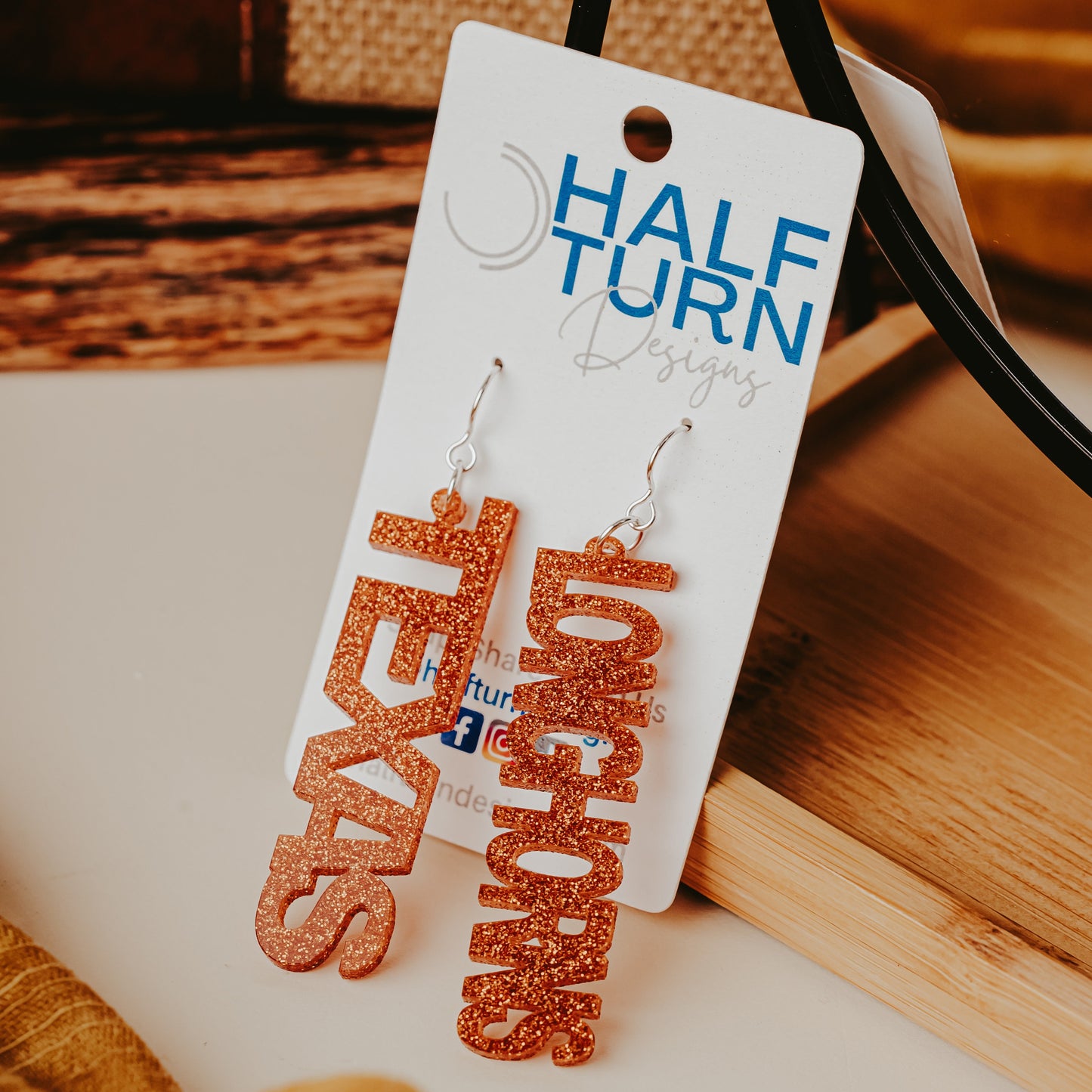 A pair of University of Texas Longhorns glitter dangle earrings on Half Turn Designs display card, featuring burnt orange acrylic word designs that spell  “TEXAS” and “LONGHORNS” on silver hooks